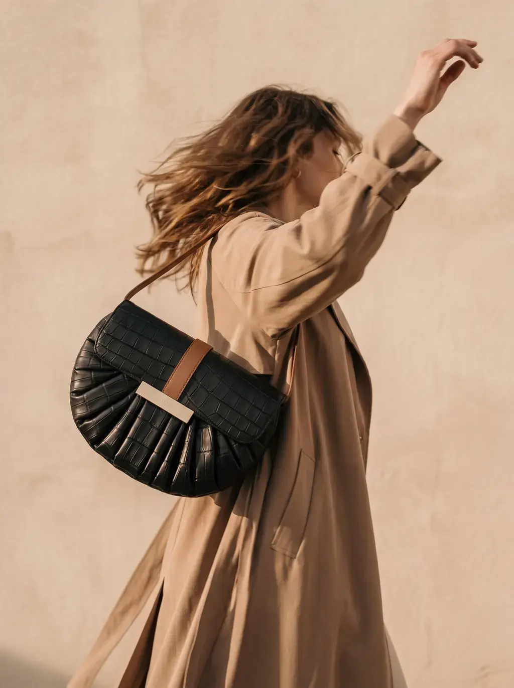 Femme en trench camel, sac croco en bandoulière, bras levés au vent