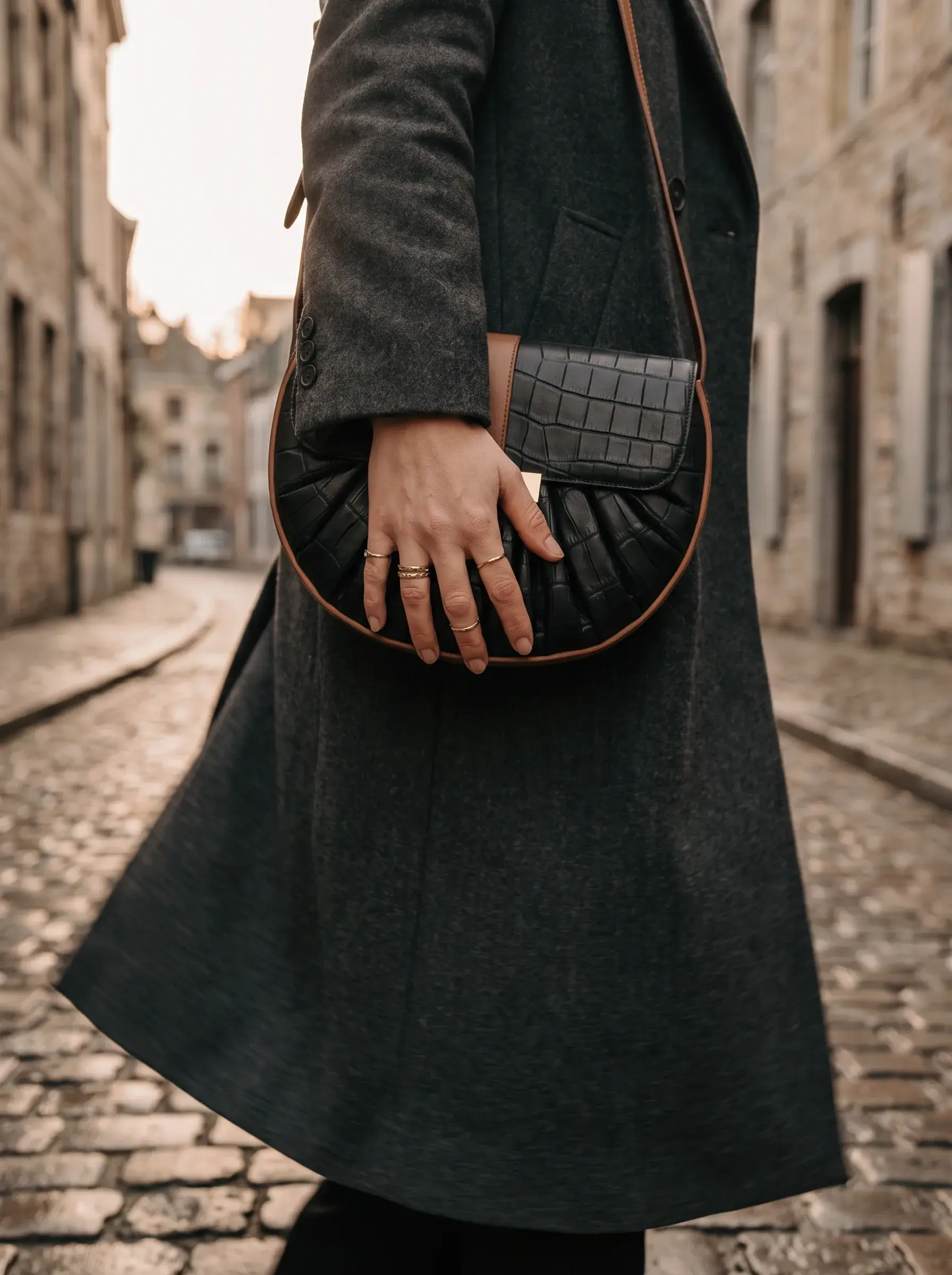 Femme en manteau noir tenant un sac tressé dans une rue pavée ancienne