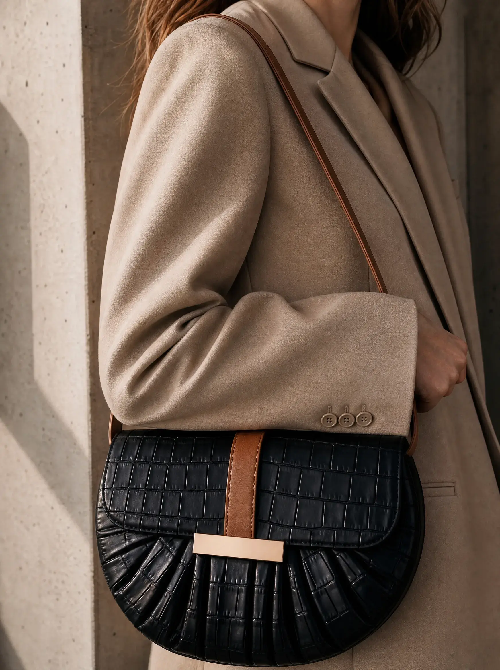 Femme en blazer beige portant un sac noir à sangle brune contre un mur clair